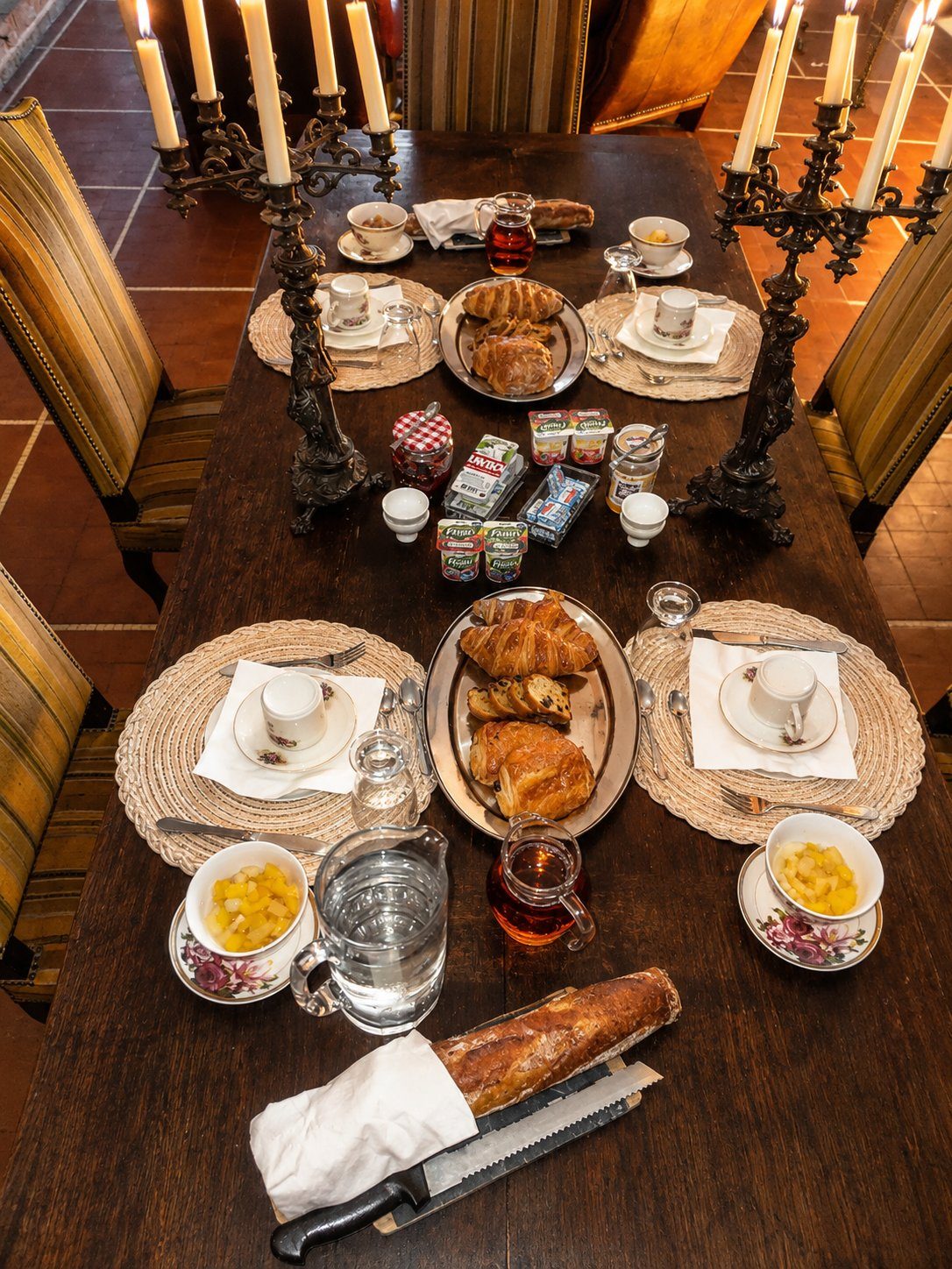 La table du petit-déjeuner dressée dans la salle à manger — viennoiseries, baguette fraîche, salade de fruits, confitures et chandeliers anciens